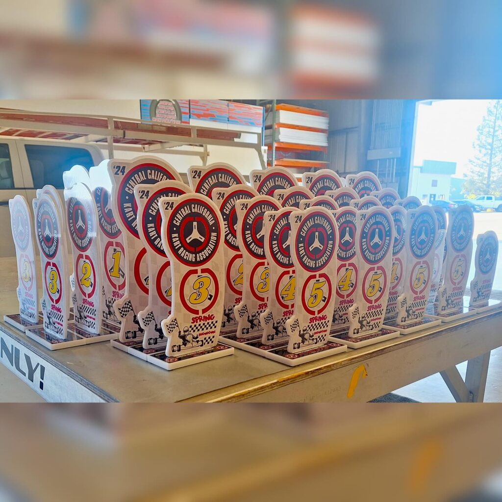 Trophies for Central California Kart Racing Association event, featuring numbered placements and a racing theme, displayed on a table in a workshop setting.
