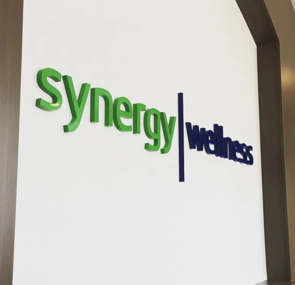 CNC-routed dimensional sign displaying "Synergy | Wellness" in green and blue lettering on a white wall, showcasing custom signage craftsmanship.