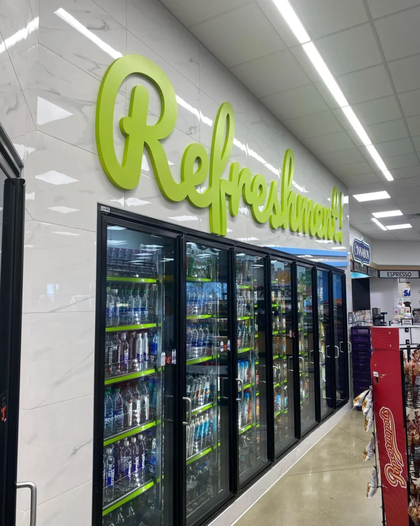 Dimensional green "Refreshment!" sign above beverage coolers in a store, showcasing various bottled drinks.