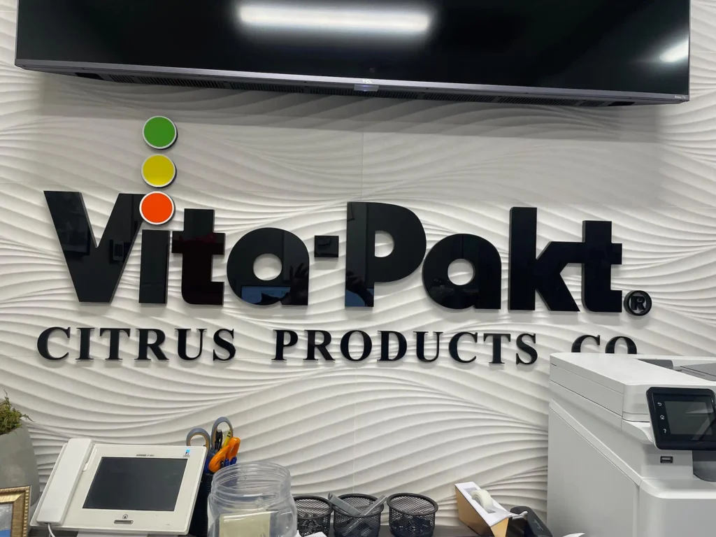 Vita-Pakt Citrus Products Co. sign displayed on textured wall, featuring colorful logo elements and surrounding office equipment.