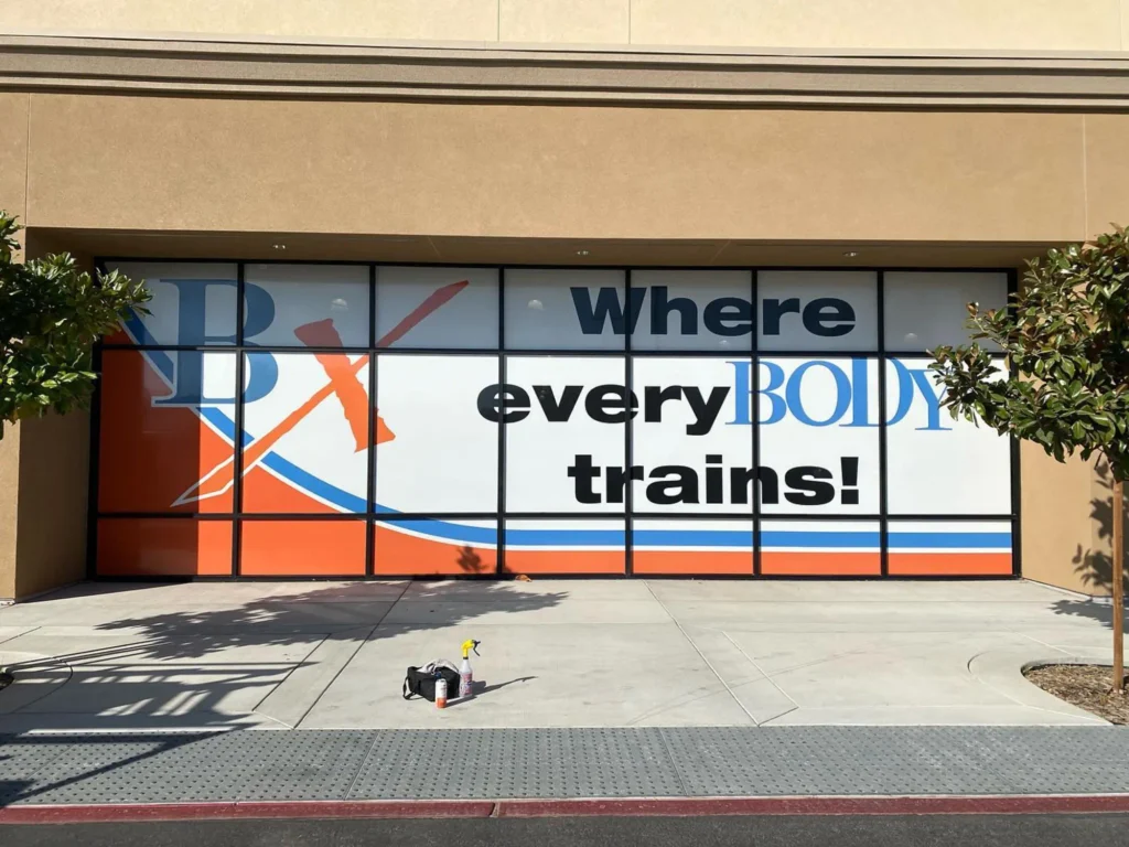 Window graphic displaying "Where every BODY trains!" with bold blue and orange design elements, showcasing branding for a fitness center.