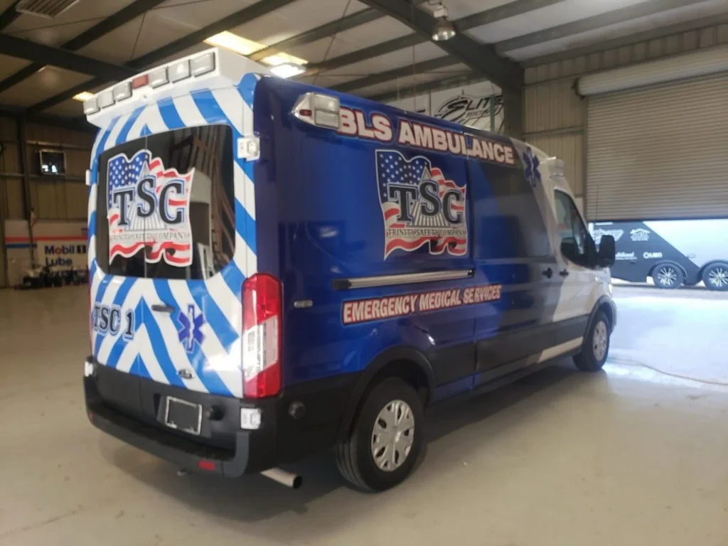Custom vehicle wrap featuring a blue and white design with "BLS Ambulance" and "TSC Emergency Medical Services" branding, displayed in a well-lit garage setting.